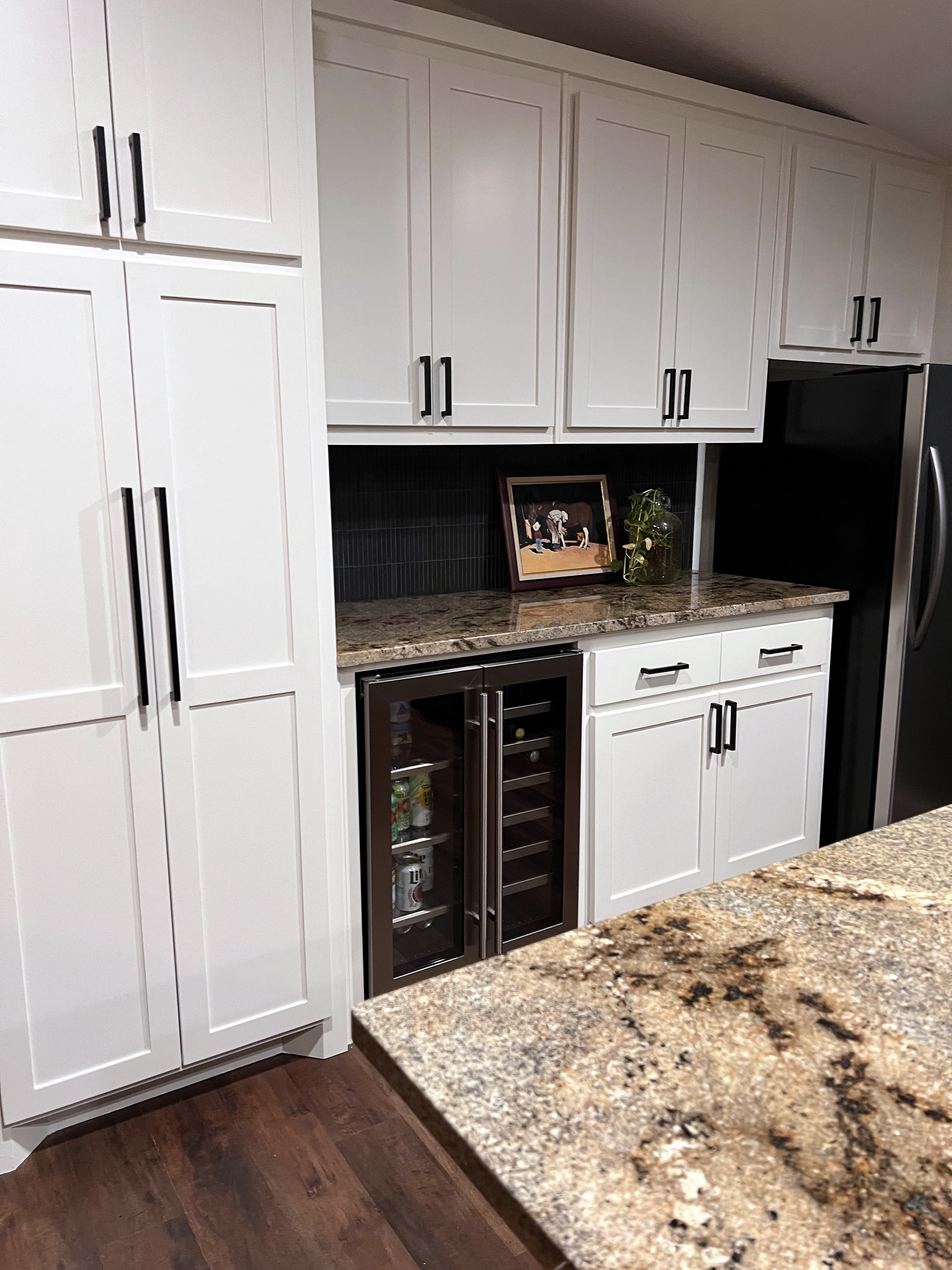 Buckhorn kitchen after — pantry tower and beverage fridge beneath white upper cabinets with black hardware, black backsplash and granite countertop