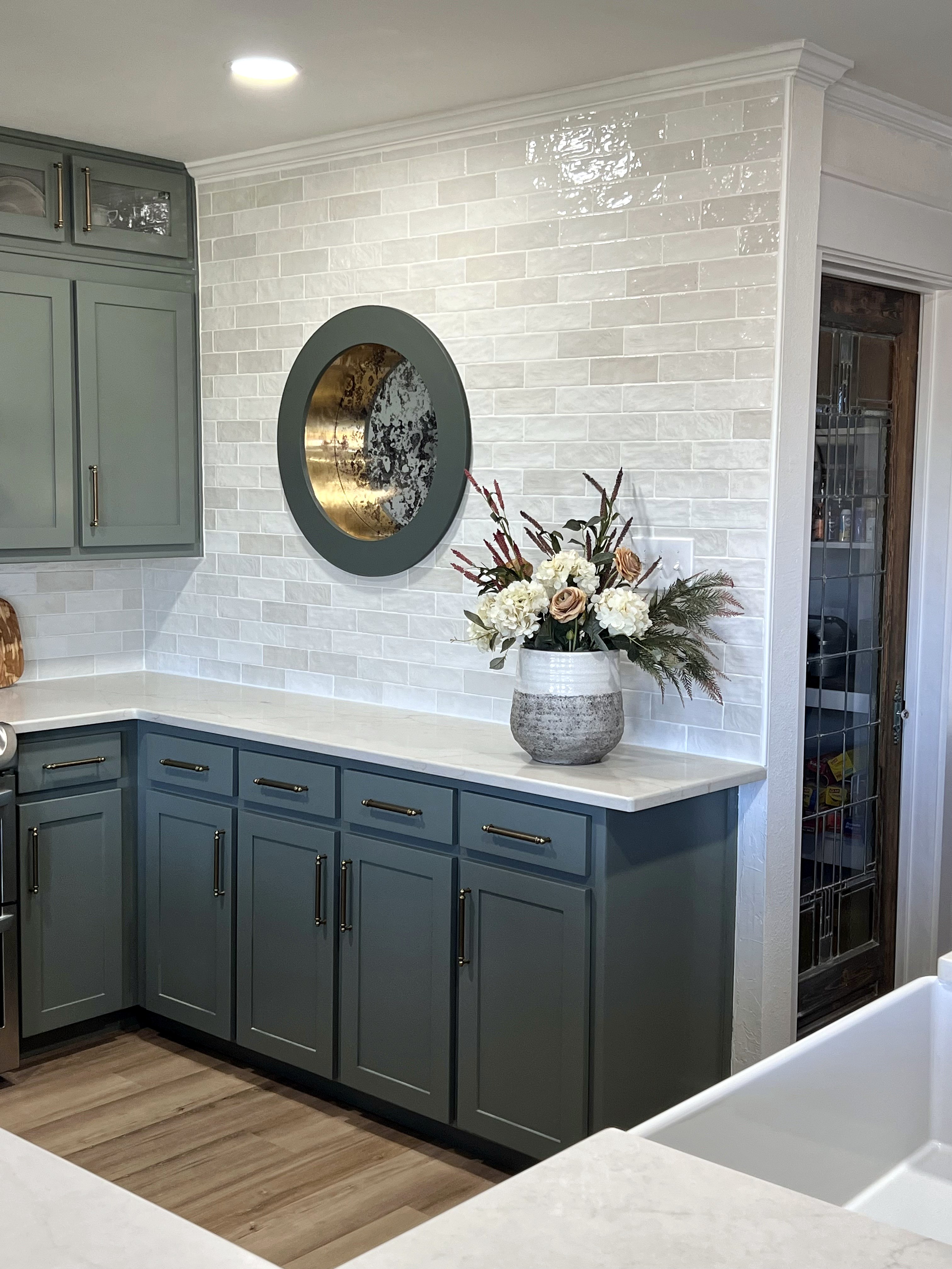 Millbrook kitchen renovation — porthole window centered on glossy subway tile backsplash wall with sage lower cabinets and flower arrangement