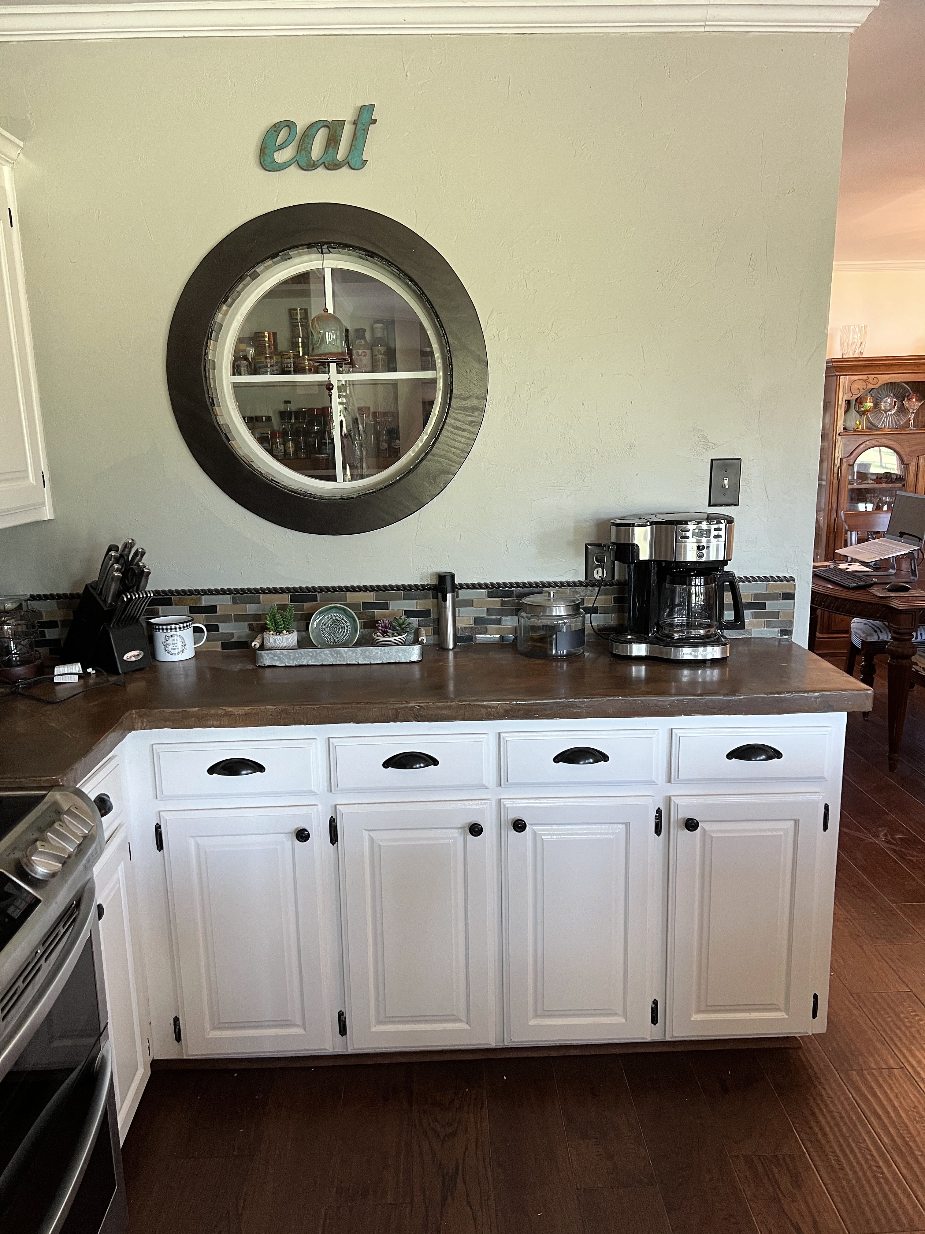 Millbrook kitchen before — original porthole window on wall with white cabinets, dark countertops and dated mosaic backsplash