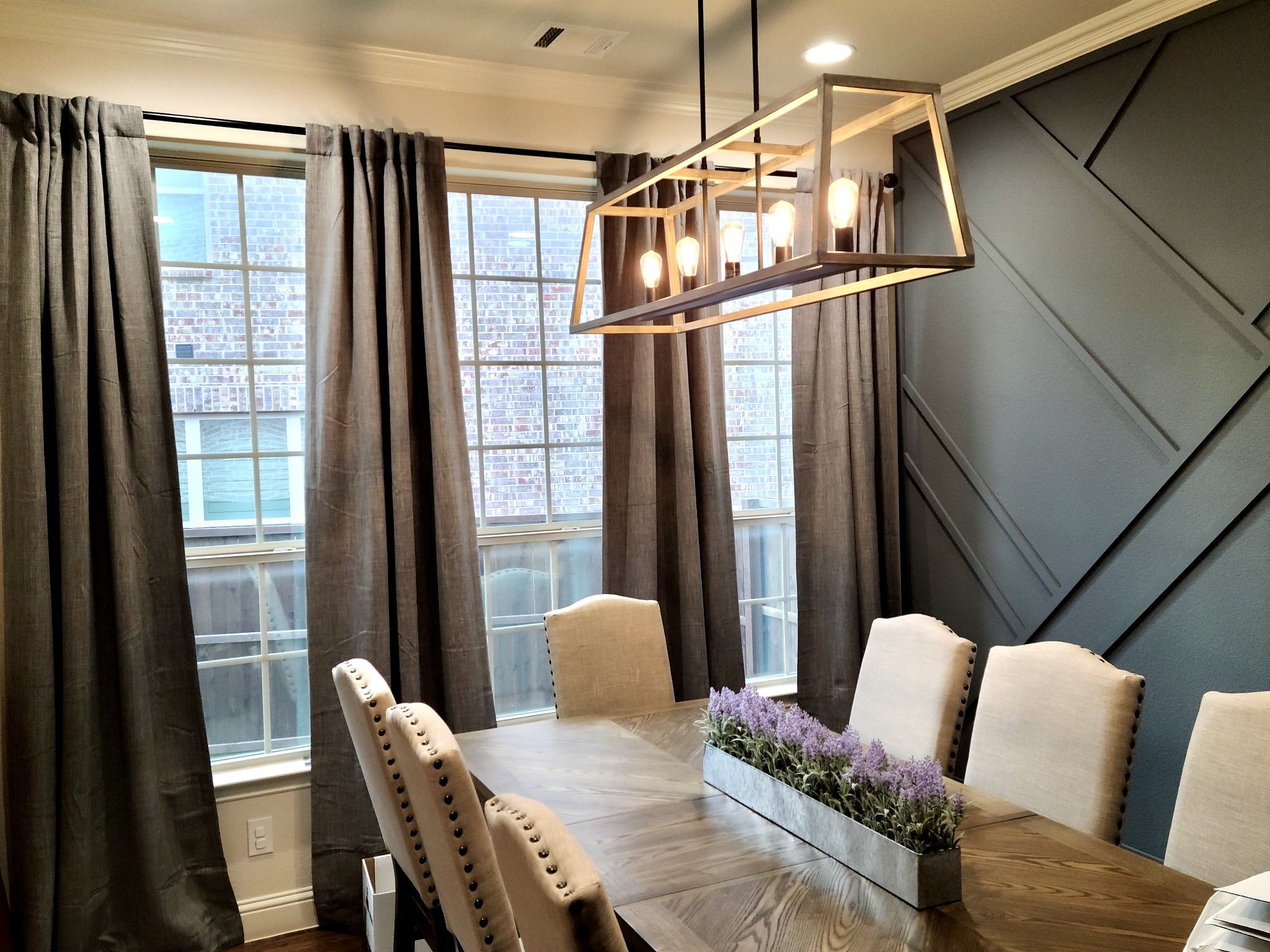 Pamela Lane dining room after — windows with gray drapery, rectangular chandelier and geometric accent wall visible above the dining table