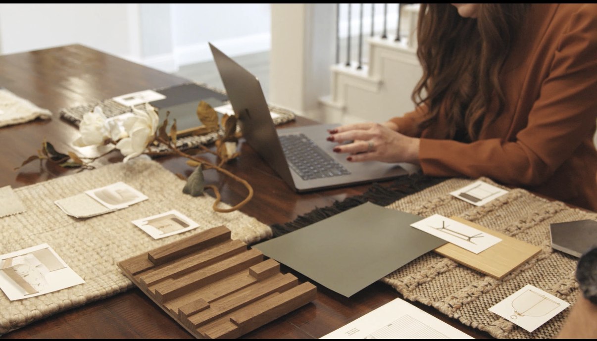 Ashley Denbow reviewing project materials, wood samples and rugs at her work table with her laptop open